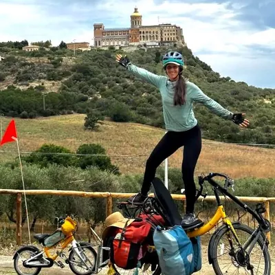 A smiling rider standing over a loaded yellow Cruzbike, arms wide, a hilltop castle behind